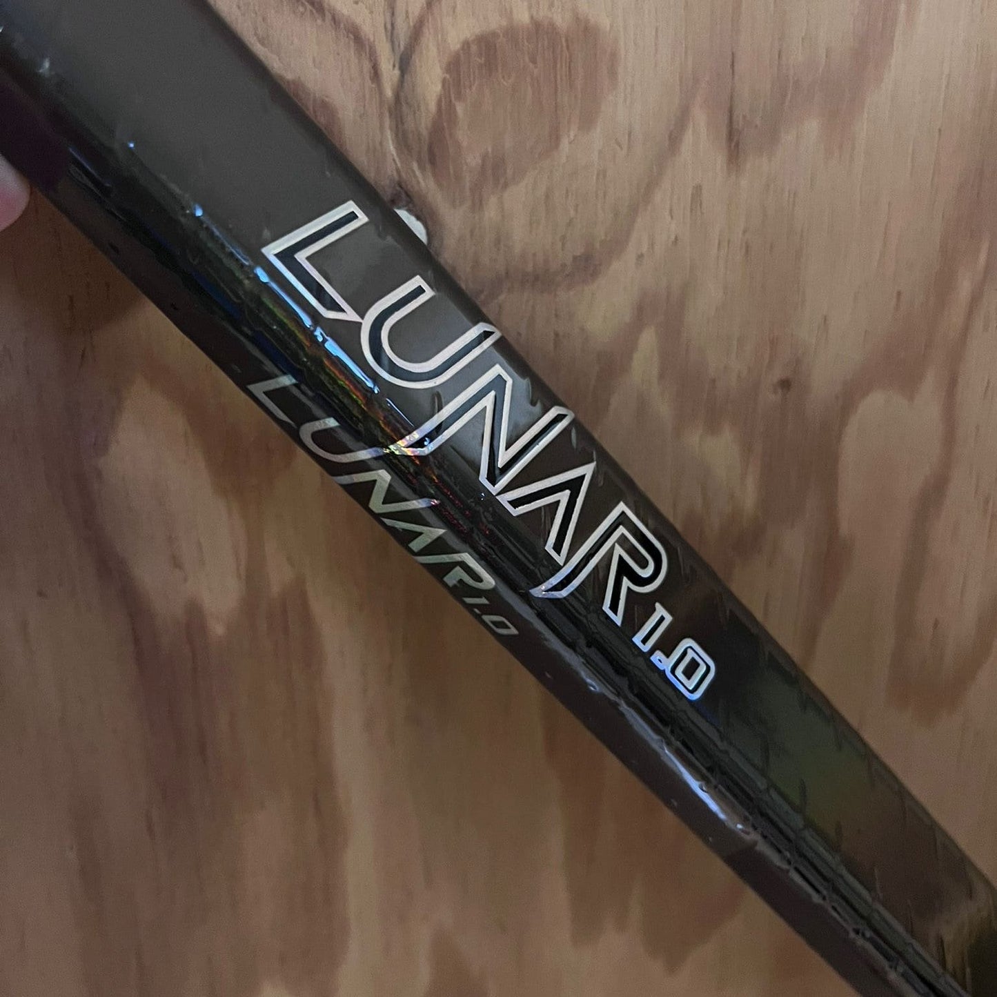 Lunar 1.0 black carbon hockey stick with subtle rainbow logo reflection on wooden background, showcasing premium quality and minimalist design.